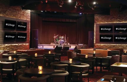 Lounge at Excalibur Hotel, a gay-friendly hotel. Stage with drum set, tables with candles, and TVs displaying the lounge logo.