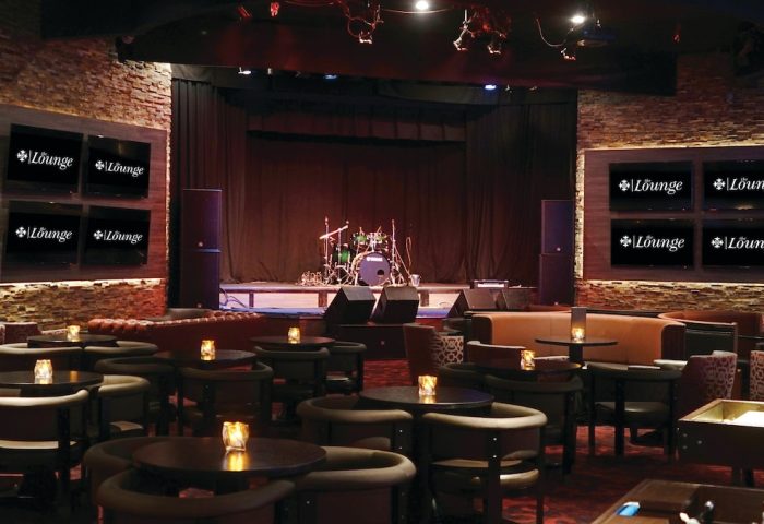 Lounge at Excalibur Hotel, a gay-friendly hotel. Stage with drum set, tables with candles, and TVs displaying the lounge logo.