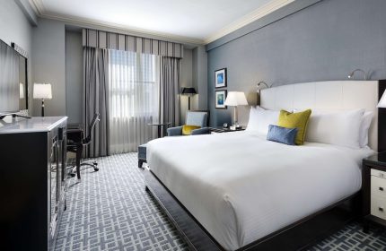 Gay-friendly Fairmont Royal York hotel room. King bed with white linens, desk, chair, and gray patterned carpet. Luxury gay travel.