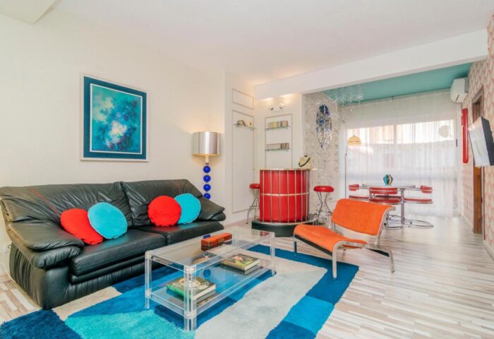 Far Home Gran Via gay-friendly hotel: Living room with black sofa, colorful pillows, bar area, and dining table. Modern design, welcoming space.