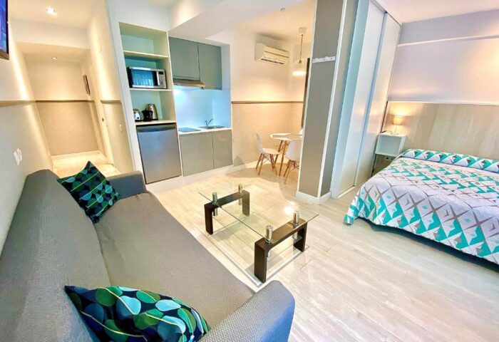 Far Home Gran Via gay-friendly hotel room. Interior view of a modern studio apartment with a sofa, bed, kitchenette, and glass table.