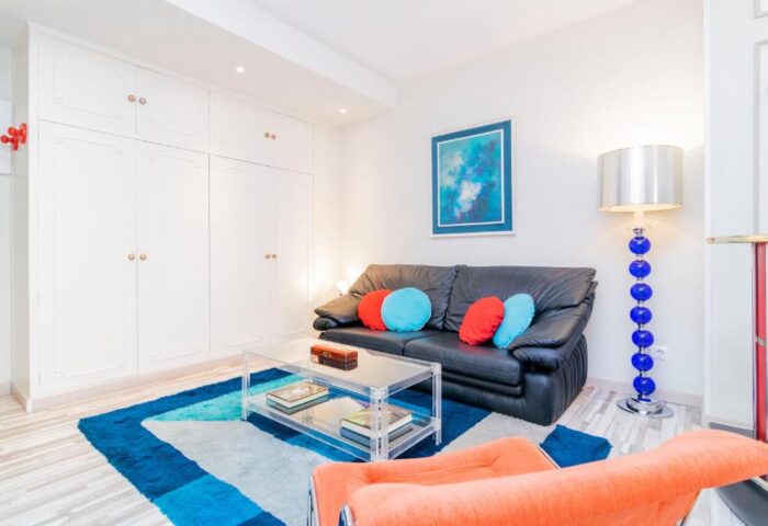 Far Home Gran Via gay-friendly hotel room. Modern interior with black sofa, colorful pillows, and a blue-themed rug and lamp. Cozy design.