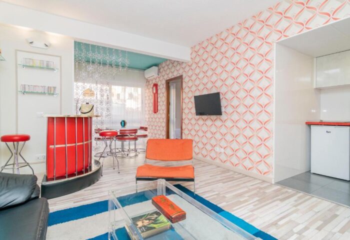 Far Home Gran Via gay-friendly hotel room. Interior with bar, stools, orange chair, TV, patterned wallpaper, and blue accents. Retro style.