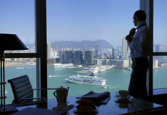 Four Seasons Hotel Hong Kong: Man adjusting tie, gazing at harbor view. Gay-friendly hotel with stunning city & water views. Upscale travel.