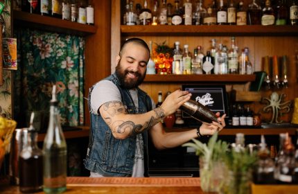 Bartender at Freehand Chicago, a gay-friendly hotel. He's shaking a cocktail. The bar is well-stocked with liquor bottles. Tattoos on his arm.