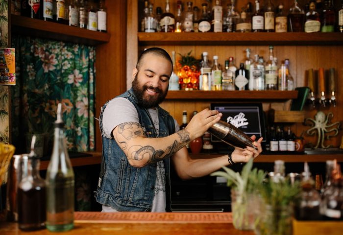 Bartender at Freehand Chicago, a gay-friendly hotel. He's shaking a cocktail. The bar is well-stocked with liquor bottles. Tattoos on his arm.