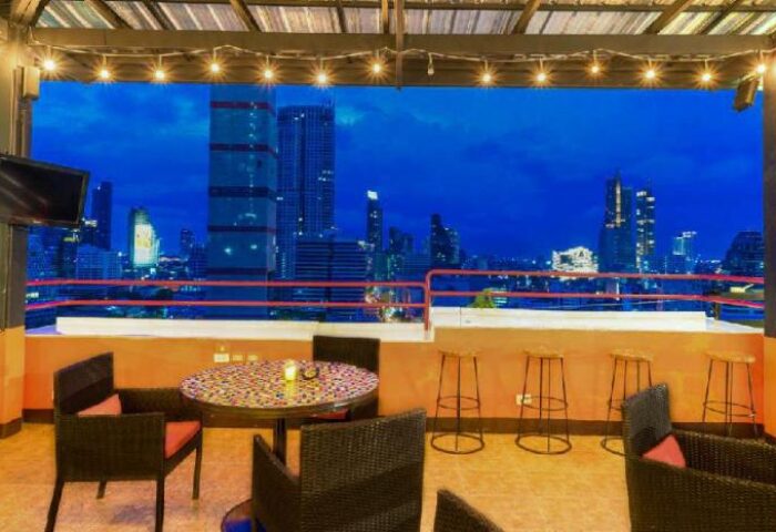 Furama Silom Hotel Bangkok rooftop bar view. Gay-friendly hotel with city skyline. Tables, chairs, and stools on the terrace at night.