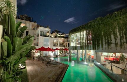 FuramaXclusive Ocean Beach Hotel Seminyak pool at night. Gay-friendly hotel with lush greenery, red umbrellas, and modern architecture.
