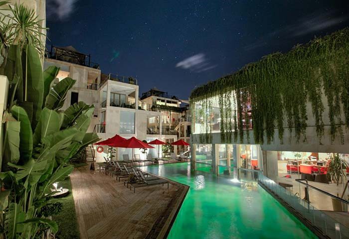 FuramaXclusive Ocean Beach Hotel Seminyak pool at night. Gay-friendly hotel with lush greenery, red umbrellas, and modern architecture.
