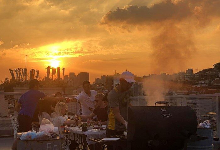 Sunset BBQ at G Guesthouse Itaewon in Seoul. Gay-friendly hotel rooftop view with men grilling, enjoying food, and the Seoul skyline.