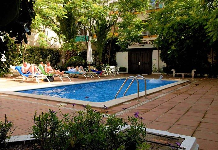 Galeon gay-friendly hotel pool. Men relax on lounge chairs by the blue pool surrounded by lush greenery. A tranquil gay travel destination.