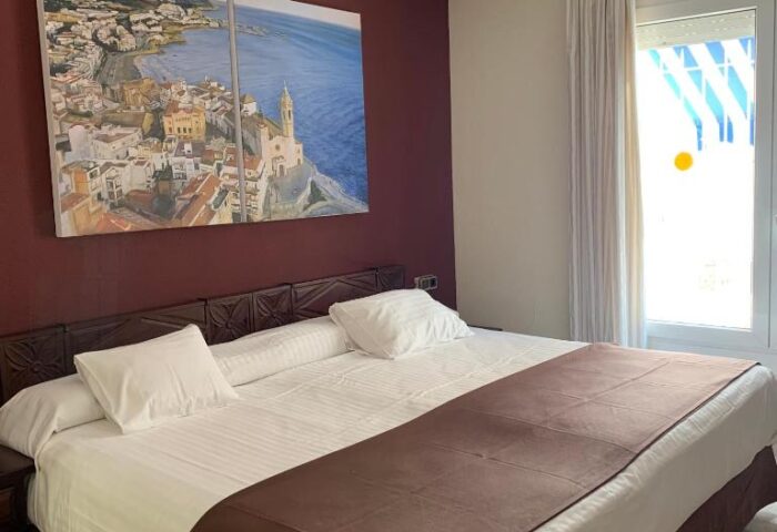 Galeon gay-friendly hotel room: double bed with pillows, brown throw, painting of a coastal town, window with curtain. Gay travel advice.