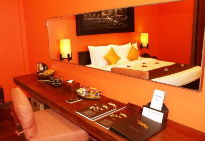 Gay-Friendly Golden Temple Hotel room. Mirror reflects bed with yellow pillows. Desk with hotel info. Orange walls. Gay travel advice.