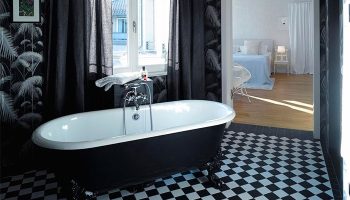Gorki Apartments' bathroom with a clawfoot tub, black and white checkered floor, palm wallpaper, and a view into the bedroom. Pet-friendly hotel.