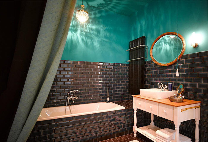 Gorki Apartments gay-friendly hotel bathroom. Dark tile, turquoise walls, vintage-style vanity, bathtub, and chandelier. Pet-friendly space.