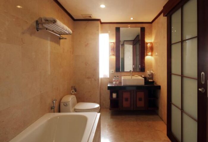 Bathroom at Grand Balisani Suites Hotel, a gay-friendly hotel. Features a bathtub, toilet, sink, mirror, and marble walls. Bali travel.
