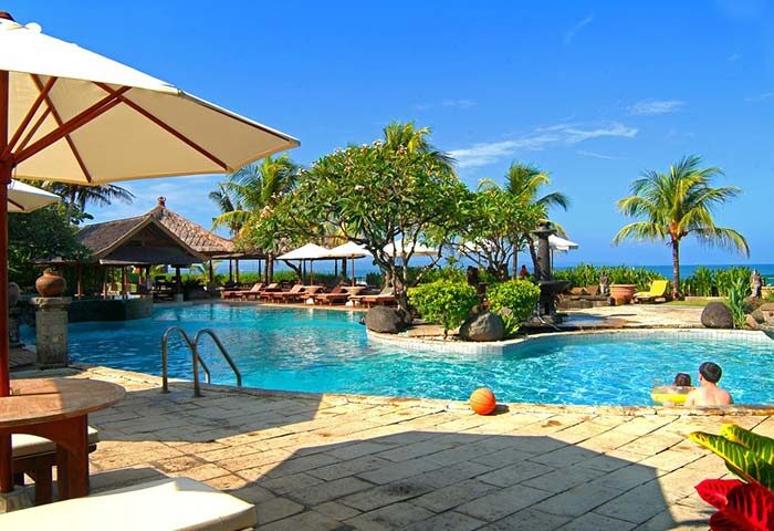Grand Balisani Suites Hotel pool view. Gay-friendly hotel with palm trees, blue sky, and clear water. Perfect for gay travel in Bali.