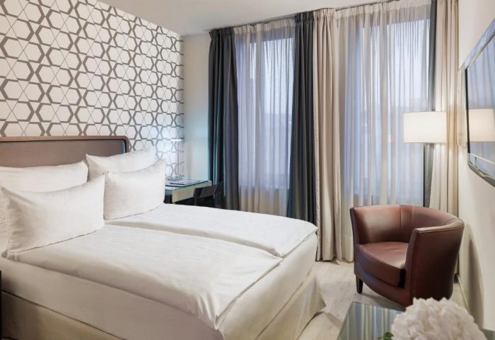 H10 Berlin Ku'damm Hotel room. Gay-friendly hotel bedroom with double bed, armchair, desk, and patterned wallpaper. Modern, comfortable design.