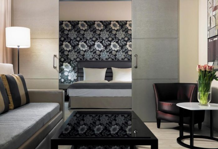 H10 Berlin Ku'damm Hotel suite. Gay-friendly hotel room with sofa, bed, floral wallpaper, and sliding doors. Modern decor.