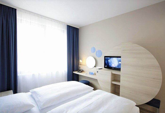 H2 Hotel Berlin-Alexanderplatz room. Modern gay-friendly hotel bedroom with twin beds, desk, TV, and blue accents. Berlin gay travel.