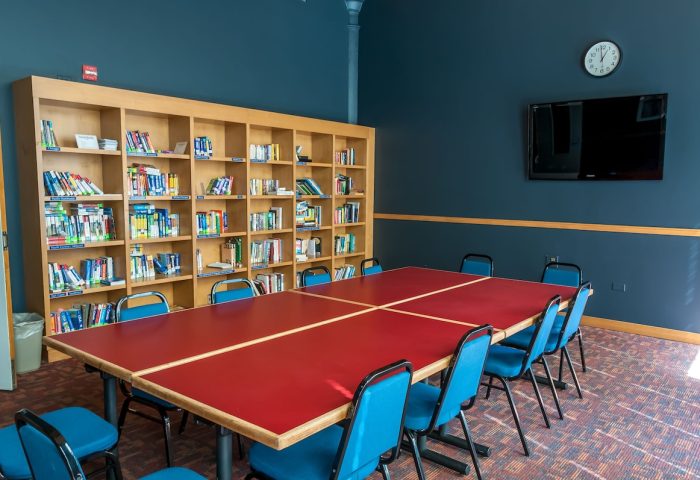 HI Chicago Hostel's common area. A large red table with blue chairs sits before a bookshelf. A TV hangs on the blue wall. Gay-friendly hotel.