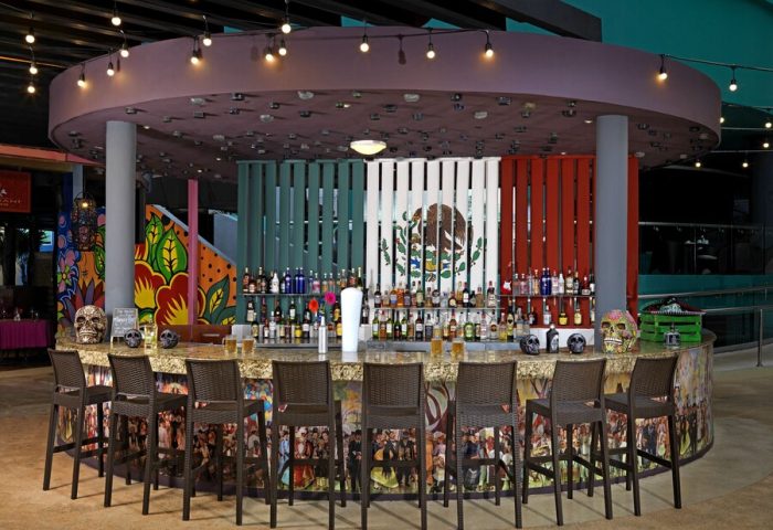 Bar at Hard Rock Hotel Cancun All Inclusive. Gay-friendly hotel with Mexican flag backdrop, skull decor, and stools. A vibrant, inclusive space.