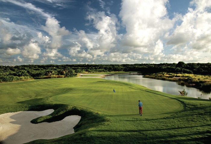 Golf course at Hard Rock Hotel Cancun All Inclusive, a gay-friendly hotel. Man on green near blue flag, water, sand trap, and trees.