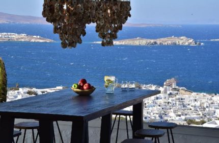 Hermes Mykonos Hotel view. Gay-friendly hotel bar table with fruit, drinks, & sea view. Mykonos town & windmills in background.