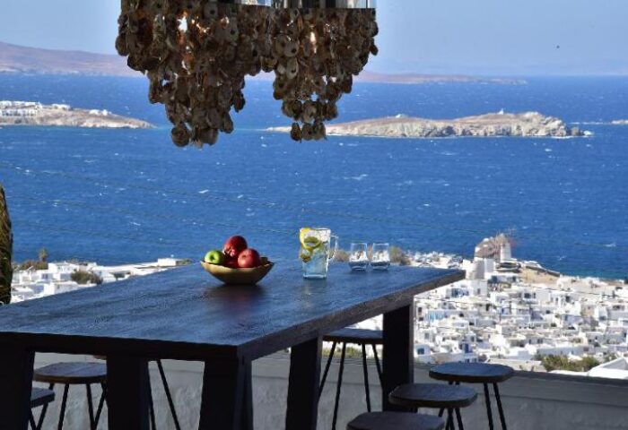 Hermes Mykonos Hotel view. Gay-friendly hotel bar table with fruit, drinks, & sea view. Mykonos town & windmills in background.