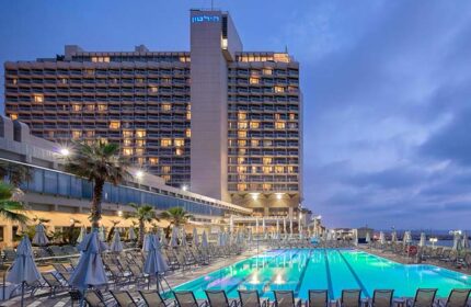 Hilton Tel Aviv, a gay-friendly hotel. View of the pool area with lounge chairs and the hotel building in the background at dusk.