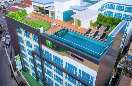 Holiday Inn Express Pattaya Central, a gay-friendly hotel. Rooftop pool, lounge chairs, tables, and greenery. Modern architecture with blue windows.