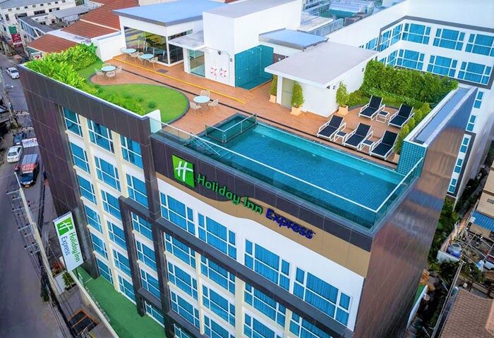 Holiday Inn Express Pattaya Central, a gay-friendly hotel. Rooftop pool, lounge chairs, tables, and greenery. Modern architecture with blue windows.
