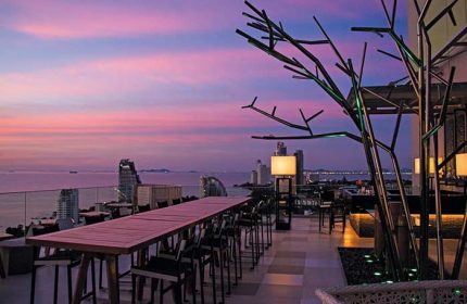 Holiday Inn Pattaya rooftop bar at sunset. Gay-friendly hotel with ocean view, tables, and decorative tree. Pattaya, Thailand.