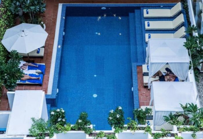 Home Chic Hotel's pool area, a gay-friendly hotel. Two men relax by the pool, one under an umbrella and another under a cabana.