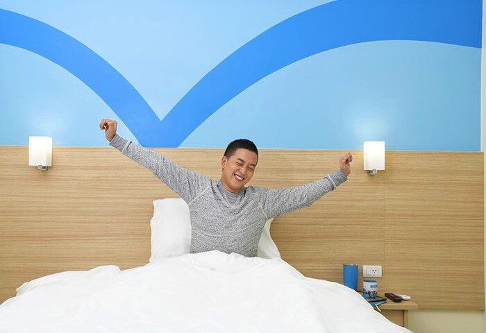 Gay-friendly Hop Inn Hotel Tomas Morato Quezon City: Man stretching in bed, smiling. Modern room, blue wall design. Comfortable stay.