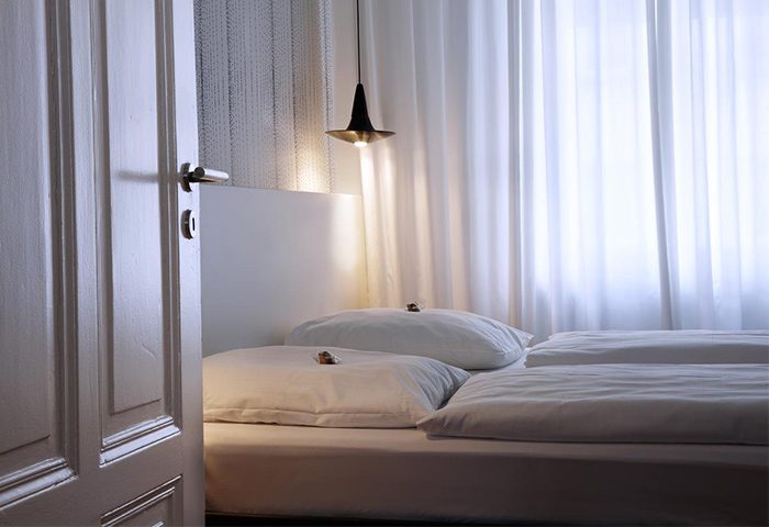 Hotel AMANO pet-friendly room with white linens, pillows, and curtains. A modern lamp hangs above the bed. Gay-friendly hotel bedroom view.