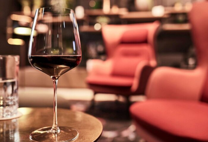 Hotel Am Konzerthaus Vienna MGallery: A glass of red wine on a table. Gay-friendly hotel lounge with red armchairs in the background.