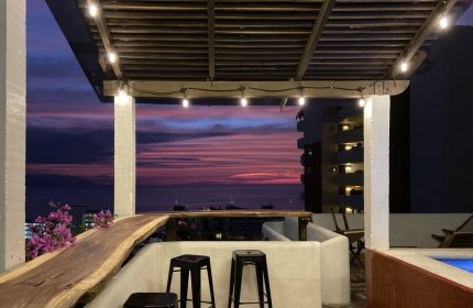 Rooftop bar at Hotel Amaca Puerto Vallarta, an adults-only, gay-friendly hotel. Sunset view with stools, lights, and ocean.