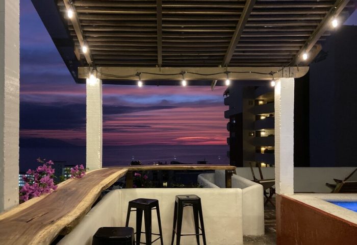 Rooftop bar at Hotel Amaca Puerto Vallarta, an adults-only, gay-friendly hotel. Sunset view with stools, lights, and ocean.