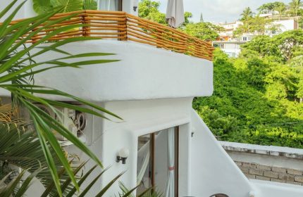 Hotel Amaca Puerto Vallarta adults-only gay-friendly hotel. Balcony view with table and chairs, lush greenery, and white building.