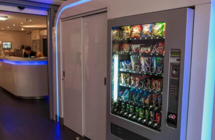 Gay-Friendly Hotel CC: Vending machine stocked with snacks and drinks. Modern design with blue lighting. Hotel reception in the background.