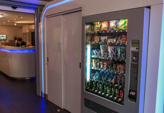 Gay-Friendly Hotel CC: Vending machine stocked with snacks and drinks. Modern design with blue lighting. Hotel reception in the background.