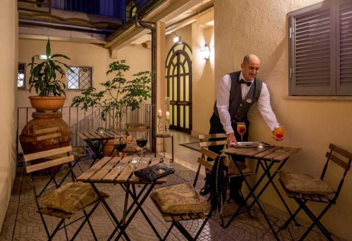 Gay-Friendly Hotel Canada, waiter serving drinks on patio. Outdoor seating with wine, cocktails, and plants. Relaxing travel experience.