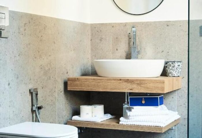 Bathroom at Gay-Friendly Hotel Central Lodge. Features floating wood shelves, round mirror, white sink, toilet, and grey tile. Modern design.