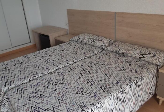 Hotel Condestable gay-friendly room. Two twin beds with patterned covers, nightstands, and a wall-mounted light fixture. Closet on the left.