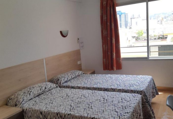 Hotel Condestable's gay-friendly room. Twin beds with patterned covers, bedside tables, and a window showing a cityscape. A comfortable stay.
