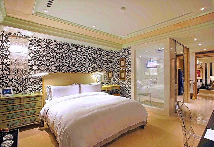 Hotel Eclat Taipei gay-friendly hotel room. Bedroom with patterned wallpaper, white bedding, and a glass-enclosed bathroom. Luxury stay.