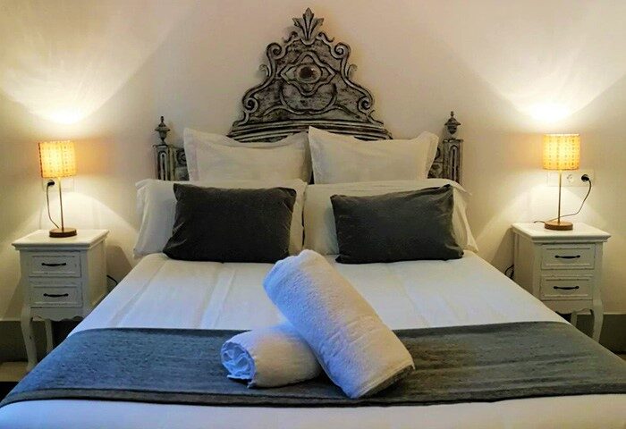 Hotel El Xalet gay-friendly room. Bed with decorative headboard, pillows, towels, bedside tables, and lamps. Gay travel lodging.