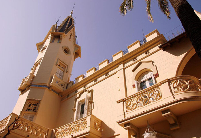 Hotel El Xalet, a gay-friendly hotel. Exterior view of the building featuring unique architecture, balconies, and a tower.