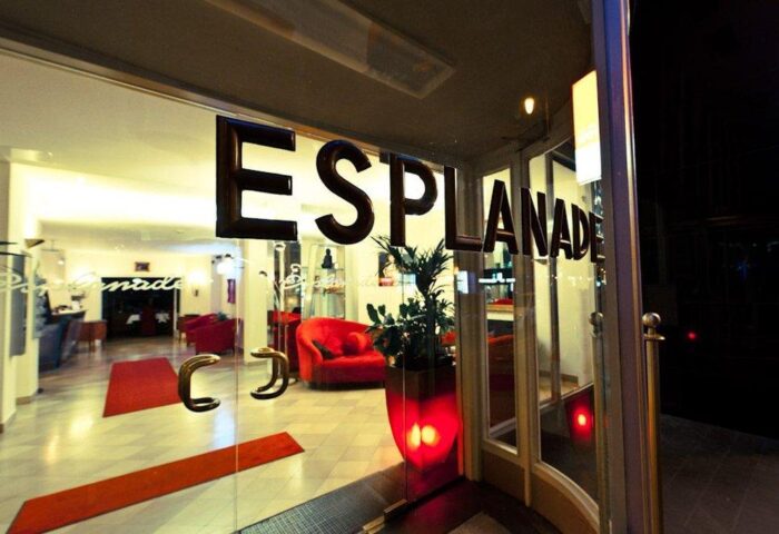 Entrance of gay-friendly Hotel Esplanade Köln. Glass door with Esplanade lettering. Red sofas and carpet visible inside. Stylish and welcoming.